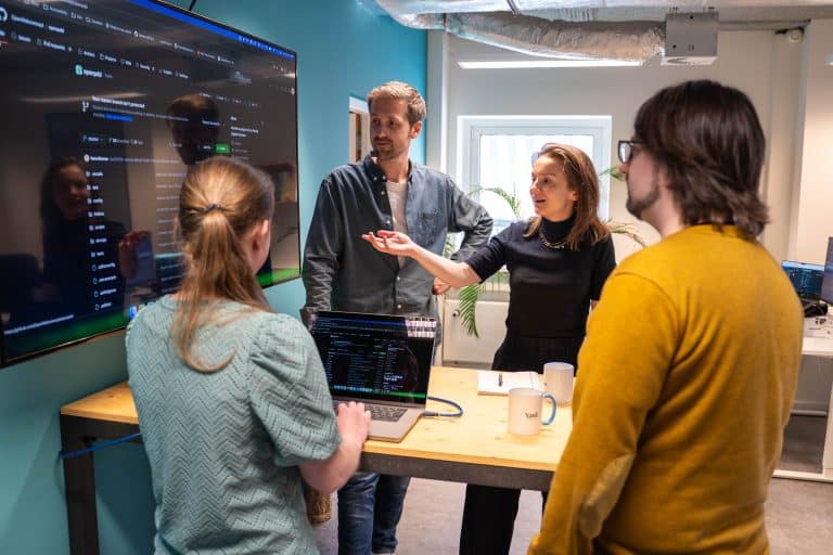 Vier mensen staan aan een hoge tafel en overleggen over iets dat ze op een scherm zien.